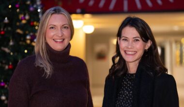 To kvinner står foran et juletre dekorert med fargerike julekuler. De befinner seg innendørs med lysende lamper som bakgrunn. En rød bakgrunn ses øverst i bildet med delvis skrift synlig. (Bildebeskrivelsen er laget av en KI-tjeneste)