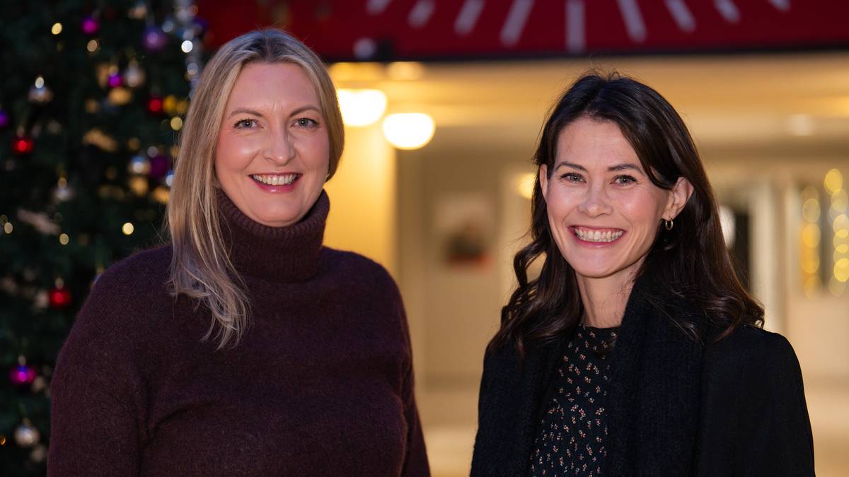 To kvinner står foran et juletre dekorert med fargerike julekuler. De befinner seg innendørs med lysende lamper som bakgrunn. En rød bakgrunn ses øverst i bildet med delvis skrift synlig. (Bildebeskrivelsen er laget av en KI-tjeneste)