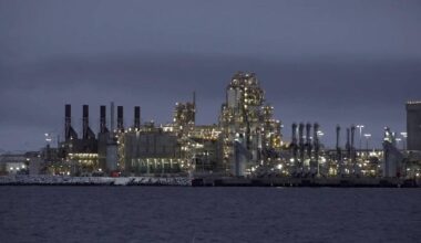 En industriverksted med mange lys og skorstener er synlig langs vannkanten. Bygningene er store med tydelige rørledninger. Omgivelsene er preget av kveldsmørke og stille vann. Aktivitet ved kaianlegget tyder på pågående drift.