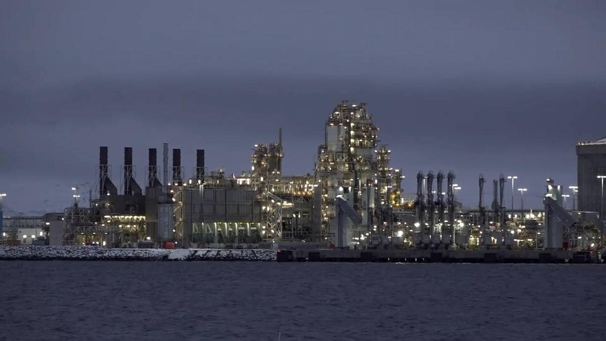 En industriverksted med mange lys og skorstener er synlig langs vannkanten. Bygningene er store med tydelige rørledninger. Omgivelsene er preget av kveldsmørke og stille vann. Aktivitet ved kaianlegget tyder på pågående drift.