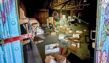 Bildet viser en rotete lagerbygning med esker og avfall spredd over gulvet. En blå dør står åpen, og en rød merkelapp med "FORSEGLING" henger fra den. I bakgrunnen er det ulike gjenstander, inkludert en sykkelhjul og verktøy. Belysningen i rommet er svakt, noe som gir en uoversiktlig atmosfære. (Bildebeskrivelsen er laget av en KI-tjeneste)