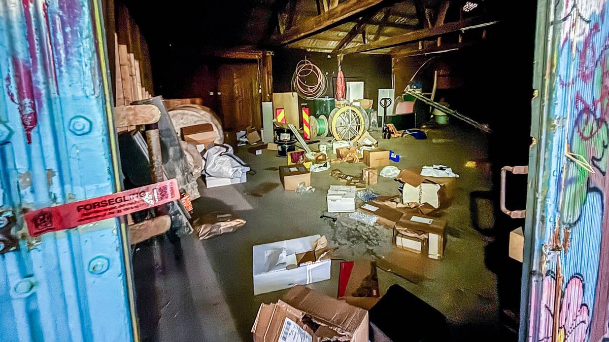 Bildet viser en rotete lagerbygning med esker og avfall spredd over gulvet. En blå dør står åpen, og en rød merkelapp med "FORSEGLING" henger fra den. I bakgrunnen er det ulike gjenstander, inkludert en sykkelhjul og verktøy. Belysningen i rommet er svakt, noe som gir en uoversiktlig atmosfære. (Bildebeskrivelsen er laget av en KI-tjeneste)