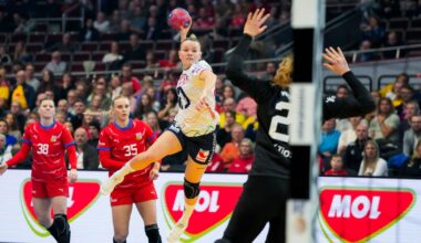 En spiller fra et håndballag hopper for å kaste ballen mot målet. To motspillere i røde drakter forsøker å blokkere kastet. Bakerst står en målvakt i svart drakt for å hindre scoring. Publikum er synlig i bakgrunnen av sportsarenaen. (Bildebeskrivelsen er laget av en KI-tjeneste)