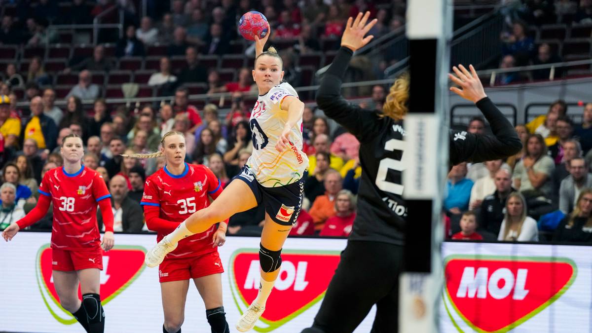 En spiller fra et håndballag hopper for å kaste ballen mot målet. To motspillere i røde drakter forsøker å blokkere kastet. Bakerst står en målvakt i svart drakt for å hindre scoring. Publikum er synlig i bakgrunnen av sportsarenaen. (Bildebeskrivelsen er laget av en KI-tjeneste)