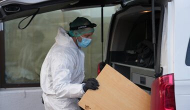 En person i hvit beskyttelsesdrakt og hansker bærer en stor trekasse fra en åpen varebil. Vedkommende har på seg en hjelm og en hette for beskyttelse. Bakgrunnen viser et vindu med tildekket glass. Omgivelsene ser ut til å være et område i bymiljø. (Bildebeskrivelsen er laget av en KI-tjeneste)