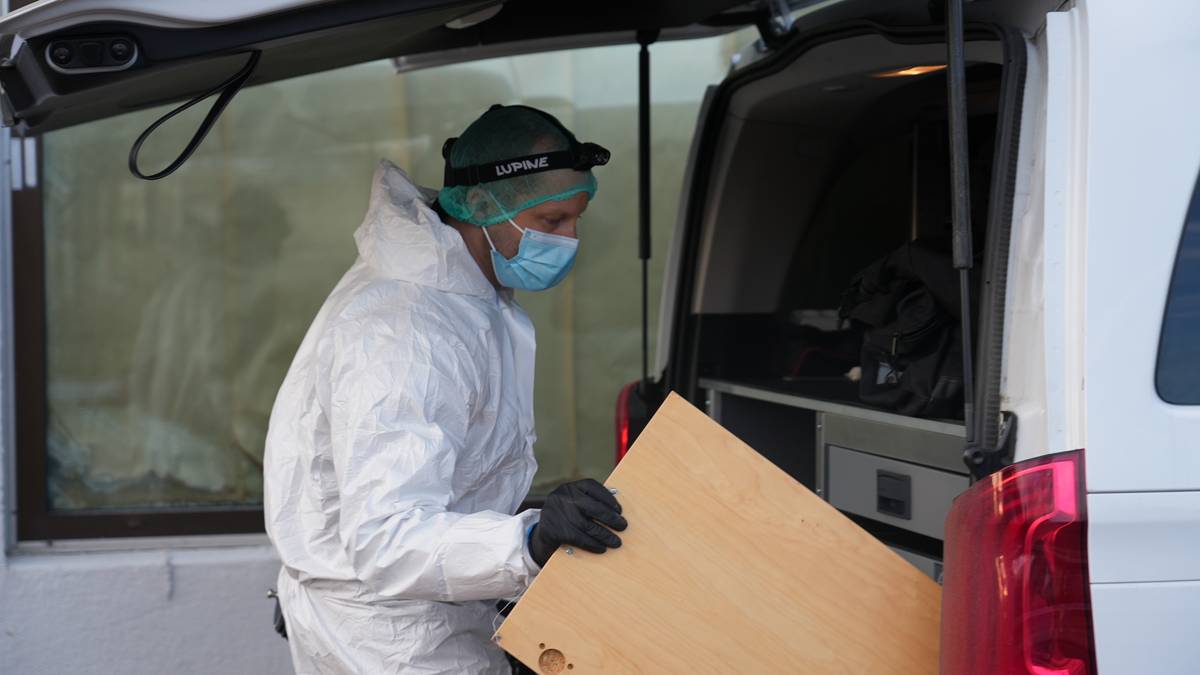 En person i hvit beskyttelsesdrakt og hansker bærer en stor trekasse fra en åpen varebil. Vedkommende har på seg en hjelm og en hette for beskyttelse. Bakgrunnen viser et vindu med tildekket glass. Omgivelsene ser ut til å være et område i bymiljø. (Bildebeskrivelsen er laget av en KI-tjeneste)