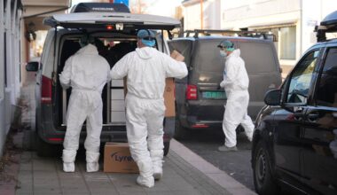 Tre personer i hvite vernedrakter laster esker inn i en varebil. Omgivelsene er en gate med flere parkerte biler. I bakgrunnen ses bygninger og fortau. Beslaget ser ut til å involvere sikkerhetsprosedyrer. (Bildebeskrivelsen er laget av en KI-tjeneste)