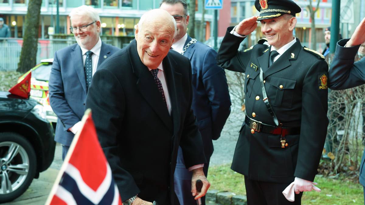 Hans Majestet Kong Harald står foran flere dresskledde personer. Han smiler. I forkant av bildet ser man et norsk flagg.