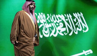 En mann ikledd tradisjonell arabisk bekledning går forbi en bakgrunn med det saudiarabiske flagget. Flagget har grønn farge med hvit skrift og et sverd. Handlingene og klesdrakten tyder på en kulturell eller offentlig sammenkomst. Omgivelsene kan indikere en offisiell seremonie eller konferanse. (Bildebeskrivelsen er laget av en KI-tjeneste)