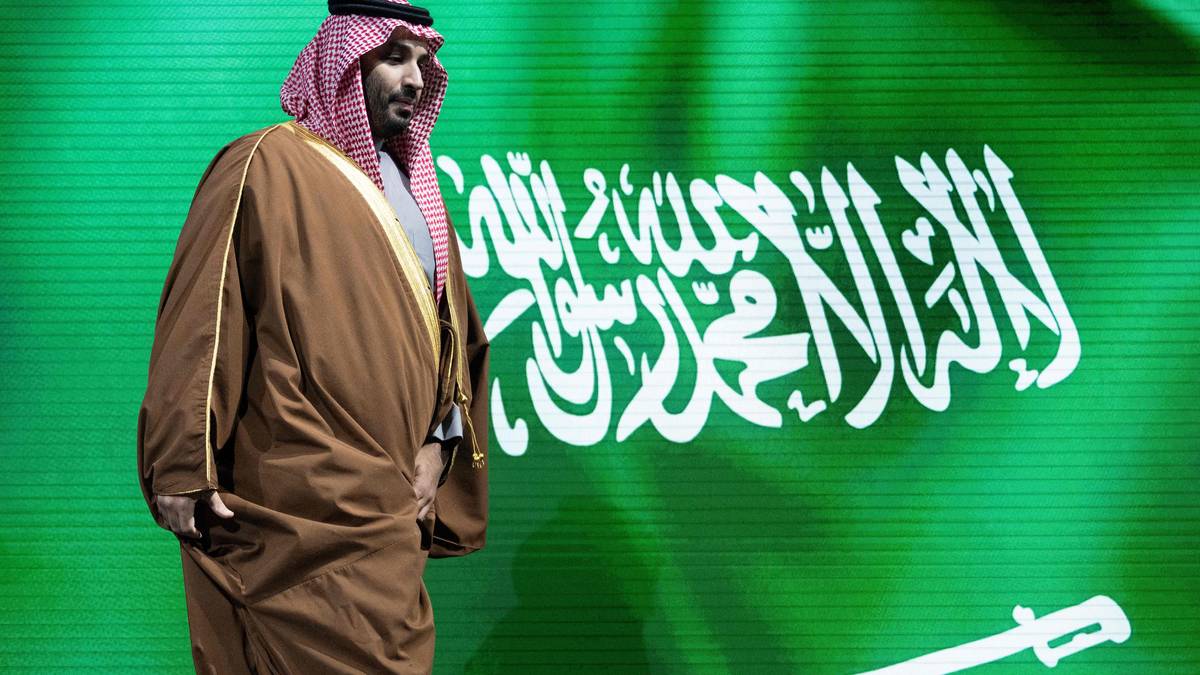 En mann ikledd tradisjonell arabisk bekledning går forbi en bakgrunn med det saudiarabiske flagget. Flagget har grønn farge med hvit skrift og et sverd. Handlingene og klesdrakten tyder på en kulturell eller offentlig sammenkomst. Omgivelsene kan indikere en offisiell seremonie eller konferanse. (Bildebeskrivelsen er laget av en KI-tjeneste)