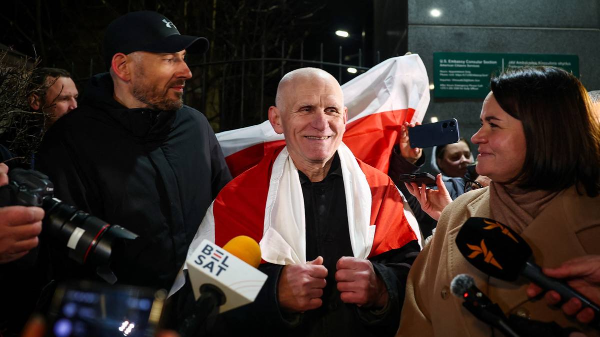 En person er dekket i et rødt og hvitt flagg, omringet av journalister med mikrofoner og kameraer. Hendelsen finner sted utenfor et bygg med grønne skilt. De tilstedeværende ser ut til å være i samtale med personen i sentrum. Flere pressemikrofoner, inkludert Belsat, er synlige i forgrunnen. (Bildebeskrivelsen er laget av en KI-tjeneste)