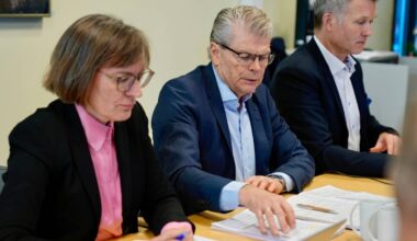 Bildet viser en gruppe personer som deltar i et møte rundt et bord. To personer er synlige i forgrunnen, en kvinne i svart jakke med rosa skjorte som holder en penn, og en mann i mørk dress som blar i et dokument. I bakgrunnen er to andre menn, en av dem kun delvis synlig. Omgivelsene inkluderer en lys kontorinnredning med datamaskiner og dokumenter på bordet. (Bildebeskrivelsen er laget av en KI-tjeneste)