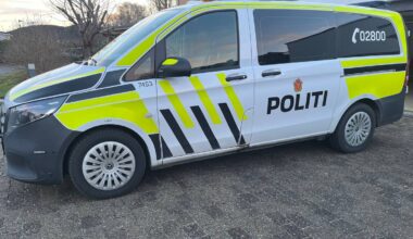 En politibil står parkert på en brosteinsbelagt gate. Bilen har tydelig merking med 'POLITI' og telefonnummeret 02800. Omgivelsene inkluderer hus og trær i bakgrunnen. Bildekomposisjonen viser venstre side av bilen. (Bildebeskrivelsen er laget av en KI-tjeneste)