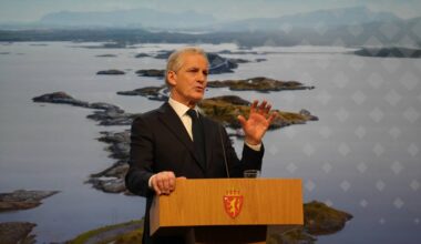 En mann står ved en talerstol med en glassvann foran seg. Bakgrunnen viser et naturskjønt kystlandskap med øyer og fjell. Mannen gestikulerer mens han er i ferd med å holde en tale. Det er lyst og klart i rommet, og han er iført en mørk dress med slips. (Bildebeskrivelsen er laget av en KI-tjeneste)