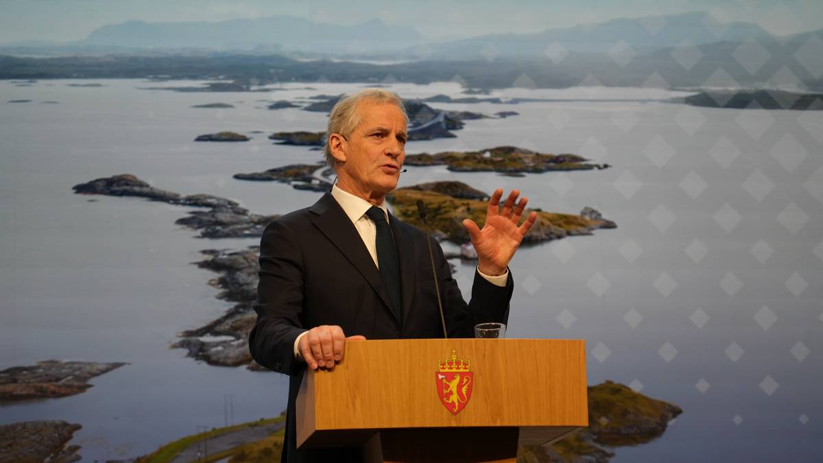 En mann står ved en talerstol med en glassvann foran seg. Bakgrunnen viser et naturskjønt kystlandskap med øyer og fjell. Mannen gestikulerer mens han er i ferd med å holde en tale. Det er lyst og klart i rommet, og han er iført en mørk dress med slips. (Bildebeskrivelsen er laget av en KI-tjeneste)