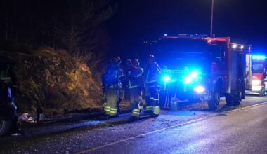 Utrykning etter en bilulykke på en mørk vei. Brannfolk står samlet ved en skadet bil og en brannbil, med blålys på. Flere biler passerer forsiktig forbi hendelsesstedet. Omgivelsene består av trær og veibelysning i bakgrunnen. (Bildebeskrivelsen er laget av en KI-tjeneste)