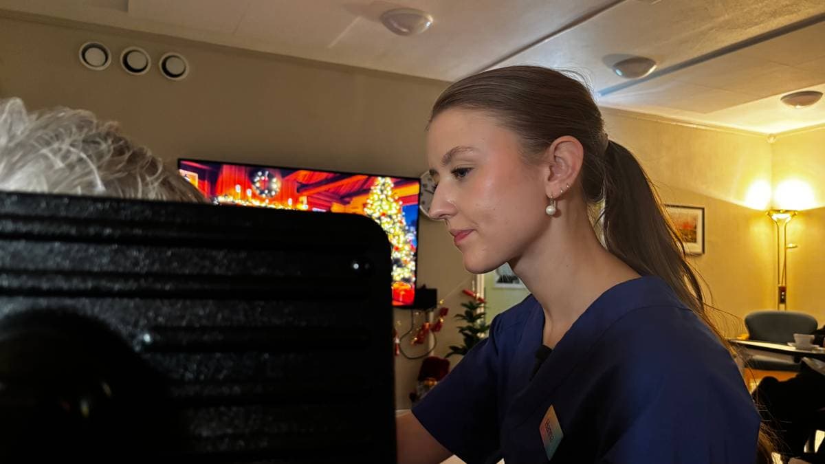 En helsearbeider i blå uniform mater en pasient. Vi ser kun nakkestøtta til rullestolen og litt av det grå håret til pasienten. I bakgrunnen er en skjerm med et juletre på.