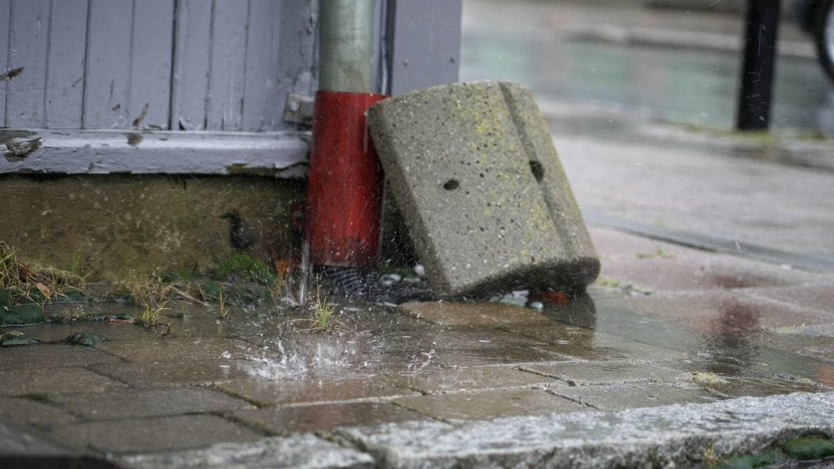 Regn vann spruter opp fra asfalt under en rørstump, med en sementblokk liggende ved foten. Omgivelsene er en bygate med våte steinheller. Det er svake grønne planter som titter frem mellom steinene. Det er overskyet vær og tydelige regndråper i luften. (Bildebeskrivelsen er laget av en KI-tjeneste)