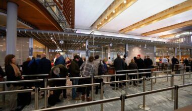 Mange mennesker står i kø ved billettkunder i en moderne terminal. Flere personer har bagasje med seg, og noen ser ned på mobiltelefonene sine. Omgivelsene har treverk og moderne interiørdetaljer. Det er lys og romslig, med synlige billettautomater og skranker.