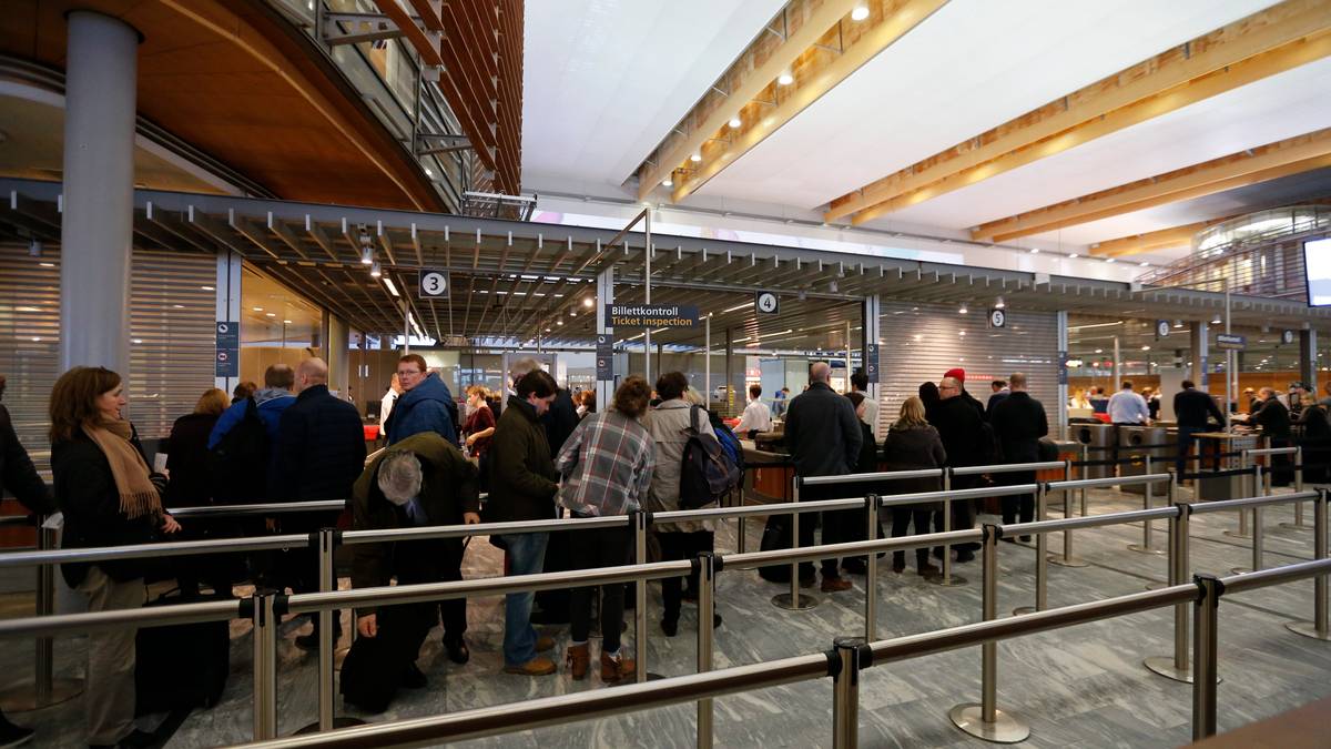 Mange mennesker står i kø ved billettkunder i en moderne terminal. Flere personer har bagasje med seg, og noen ser ned på mobiltelefonene sine. Omgivelsene har treverk og moderne interiørdetaljer. Det er lys og romslig, med synlige billettautomater og skranker.
