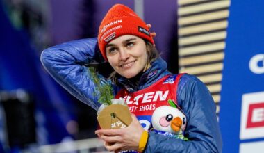 En utøver står på seierspallen iført en rød lue og en blå jakke. Vedkommende holder en pokal med grønne kvister og en kosedyrfigur i hånden. Bakgrunnen viser en konkurransearena med en større plakat. Det er kveldsbelysning på stedet, som gir en atmosfære av feiring. (Bildebeskrivelsen er laget av en KI-tjeneste)