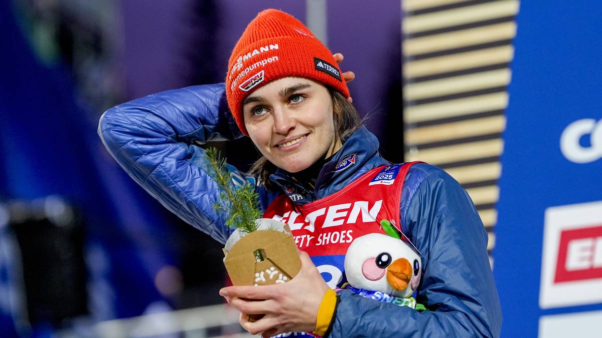 En utøver står på seierspallen iført en rød lue og en blå jakke. Vedkommende holder en pokal med grønne kvister og en kosedyrfigur i hånden. Bakgrunnen viser en konkurransearena med en større plakat. Det er kveldsbelysning på stedet, som gir en atmosfære av feiring. (Bildebeskrivelsen er laget av en KI-tjeneste)