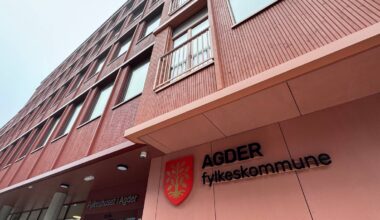 Fylkeshuset i Agder fotografert fra bakkenivå med fokus på bygningens fasade. Bygget har røde vegger og flere vinduer. Skiltet med teksten "AGDER fylkeskommune" er synlig ved inngangen. Omgivelsene er grå og skyet, noe som gir et dyster preg. (Bildebeskrivelsen er laget av en KI-tjeneste)