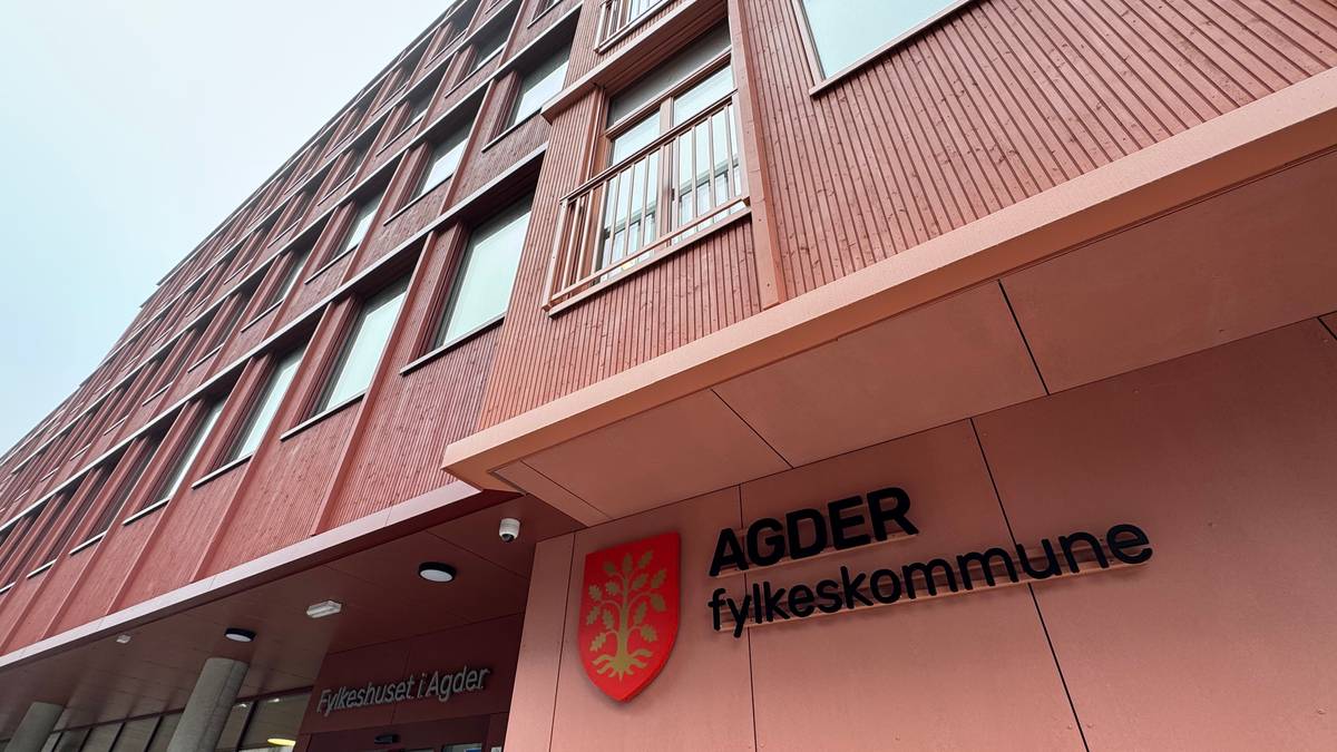 Fylkeshuset i Agder fotografert fra bakkenivå med fokus på bygningens fasade. Bygget har røde vegger og flere vinduer. Skiltet med teksten "AGDER fylkeskommune" er synlig ved inngangen. Omgivelsene er grå og skyet, noe som gir et dyster preg. (Bildebeskrivelsen er laget av en KI-tjeneste)