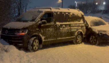 To biler står parkert i snøen, en mørk Volkswagen varebil og en annen bil delvis dekket av snø. Omgivelsene er snødekte, og det er mørkt. Bilene har snø på tak og vinduer, og snøen er synlig på bakken rundt dem. Det er ingen personer synlige i bildet.