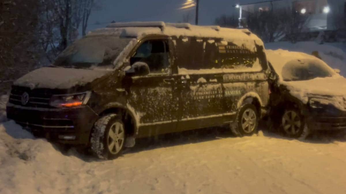 To biler står parkert i snøen, en mørk Volkswagen varebil og en annen bil delvis dekket av snø. Omgivelsene er snødekte, og det er mørkt. Bilene har snø på tak og vinduer, og snøen er synlig på bakken rundt dem. Det er ingen personer synlige i bildet.