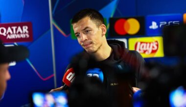 En sportsutøver fra FK Bodø/Glimt står foran medier under en pressekonferanse. Han holder en mikrofon og svarer på spørsmål fra journalistene. I bakgrunnen er reklameplakater for sponsorer synlige. Det er flere kamerautstyr og mikrofoner i nærheten, som indikerer en travelt mediedekning. (Bildebeskrivelsen er laget av en KI-tjeneste)