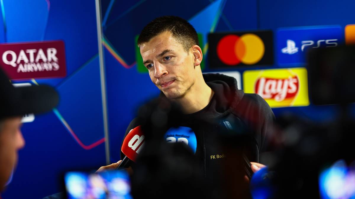 En sportsutøver fra FK Bodø/Glimt står foran medier under en pressekonferanse. Han holder en mikrofon og svarer på spørsmål fra journalistene. I bakgrunnen er reklameplakater for sponsorer synlige. Det er flere kamerautstyr og mikrofoner i nærheten, som indikerer en travelt mediedekning. (Bildebeskrivelsen er laget av en KI-tjeneste)