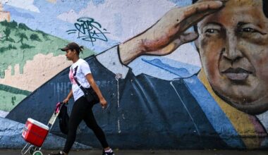 En kvinne går forbi en fargerik veggmaleri mens hun drar på en rød vogn. Hun har på seg en t-skjorte med trykk og joggesko. Omgivelsene viser en urban bakgrunn med graffiti på muren. Det er dag, og belysningen er naturlig. (Bildebeskrivelsen er laget av en KI-tjeneste)
