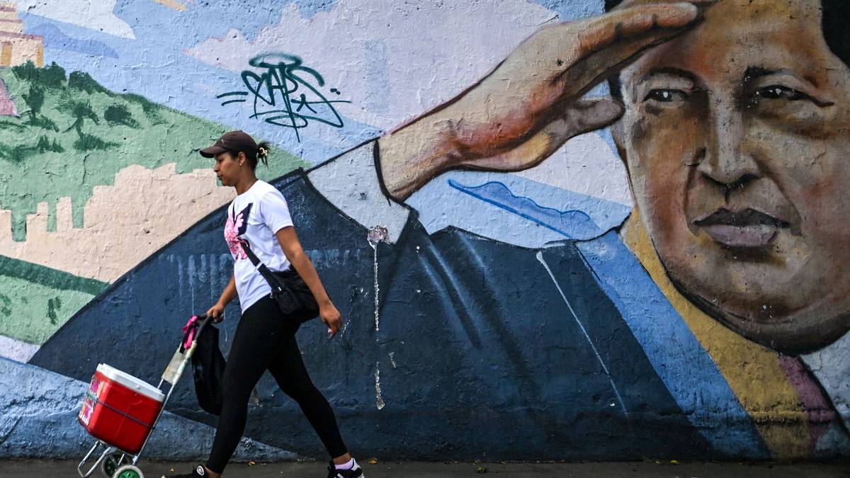 En kvinne går forbi en fargerik veggmaleri mens hun drar på en rød vogn. Hun har på seg en t-skjorte med trykk og joggesko. Omgivelsene viser en urban bakgrunn med graffiti på muren. Det er dag, og belysningen er naturlig. (Bildebeskrivelsen er laget av en KI-tjeneste)
