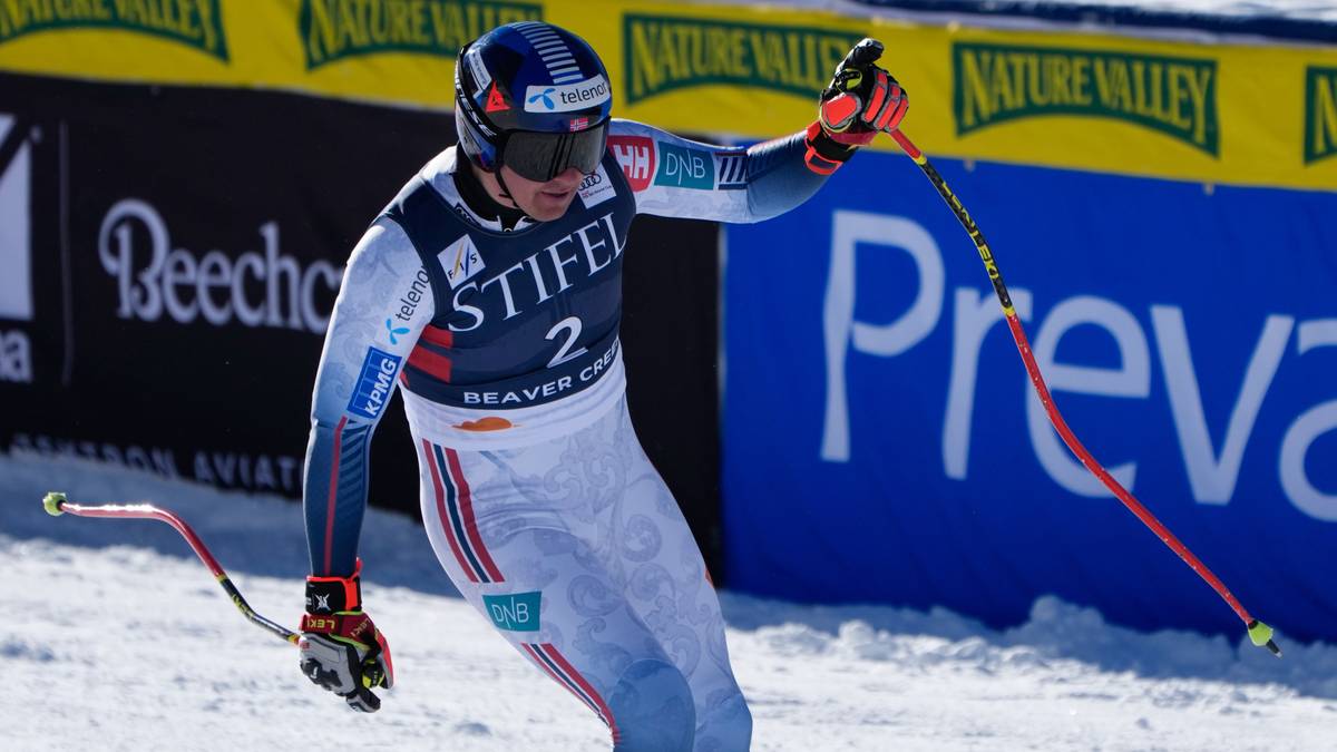 En alpinist iført konkurransekledd med nummer 2 fører håndflatene opp og feirer. Bakgrunnen viser snødekte bakker og reklameplakater. Utstyr som ski og staver er synlige i bildet. Det er klart solfylt vær.