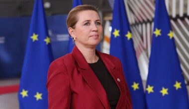 En kvinne står foran flere EU-flagg i en offisiell setting. Hun har på seg en rød blazer og blikket rettet fremover. Bakgrunnen viser et moderne rom, delvis opptatt av et rødt teppe. Bildekomposisjonen signaliserer et formelt arrangement. (Bildebeskrivelsen er laget av en KI-tjeneste)