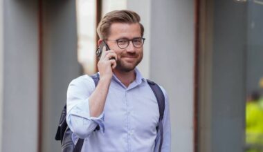 En mann iført en lys skjorte og med en skulderveske går langs en gate mens han snakker i mobiltelefon. Det er en urban setting med bygninger i bakgrunnen. Mannen bruker også et headset. Omgivelsene tyder på et travelt miljø. (Bildebeskrivelsen er laget av en KI-tjeneste)