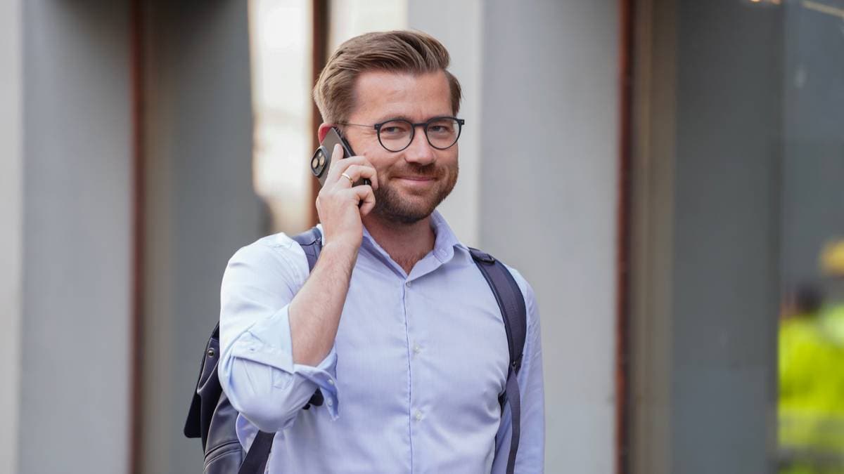 En mann iført en lys skjorte og med en skulderveske går langs en gate mens han snakker i mobiltelefon. Det er en urban setting med bygninger i bakgrunnen. Mannen bruker også et headset. Omgivelsene tyder på et travelt miljø. (Bildebeskrivelsen er laget av en KI-tjeneste)