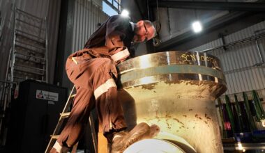 En arbeidstaker står på en stige og arbeider med en stor metallkomponent i et industrielt miljø. Lys fra taklamper belyser området, og det er en stålstige synlig i bakgrunnen. Både arbeidsklærne og omgivelsene er preget av bruk. Det er også en skuff med fargede materialer hengende på veggen.