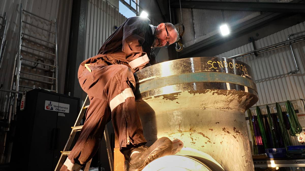 En arbeidstaker står på en stige og arbeider med en stor metallkomponent i et industrielt miljø. Lys fra taklamper belyser området, og det er en stålstige synlig i bakgrunnen. Både arbeidsklærne og omgivelsene er preget av bruk. Det er også en skuff med fargede materialer hengende på veggen.