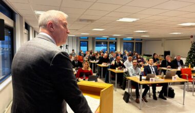 Et møterom med mange deltakere som sitter ved bord med datamaskiner, drikkevarer og notater. Foran i bildet står en person ved en talerstol, som henvender seg til publikum. Under møtet er det forskjellige oppsett av stoler, og et juletre er synlig i bakgrunnen. Rommet har store vinduer som slipper inn naturlig lys. (Bildebeskrivelsen er laget av en KI-tjeneste)