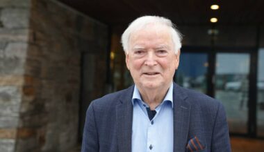 En eldre mann står foran en bygning med steinvegg og glassdør. Han har på seg en mørk blazer og lyse skjorte. Bakgrunnen viser gråstein og klar belysning. Atmosfæren er nøytral og informativ. (Bildebeskrivelsen er laget av en KI-tjeneste)