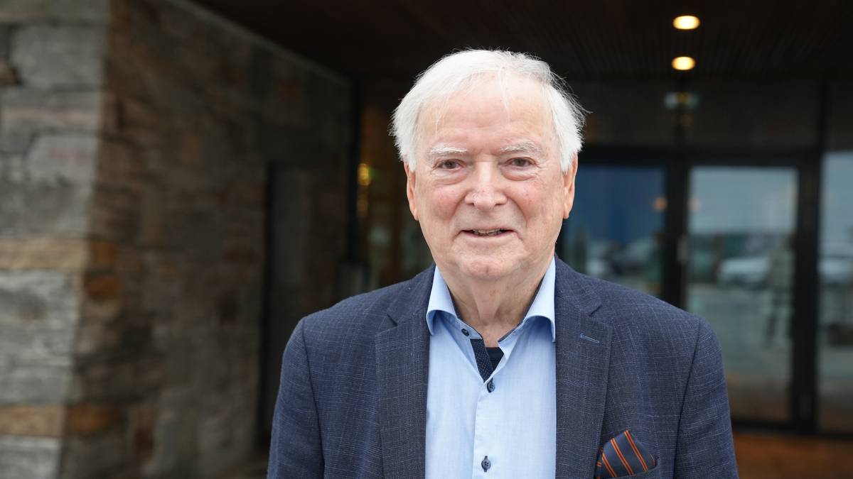 En eldre mann står foran en bygning med steinvegg og glassdør. Han har på seg en mørk blazer og lyse skjorte. Bakgrunnen viser gråstein og klar belysning. Atmosfæren er nøytral og informativ. (Bildebeskrivelsen er laget av en KI-tjeneste)