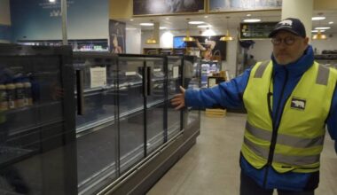 En ansatt i en butikk står foran tomme kjøleskapseksjoner og gestikulerer med hånden. Butikken har en moderne innredning med belysning og hyller i bakgrunnen. Det er flere produkter synlige i de andre kjøleskapene til venstre. En plakat på veggen med bilder av dyr kan sees i bakgrunnen.