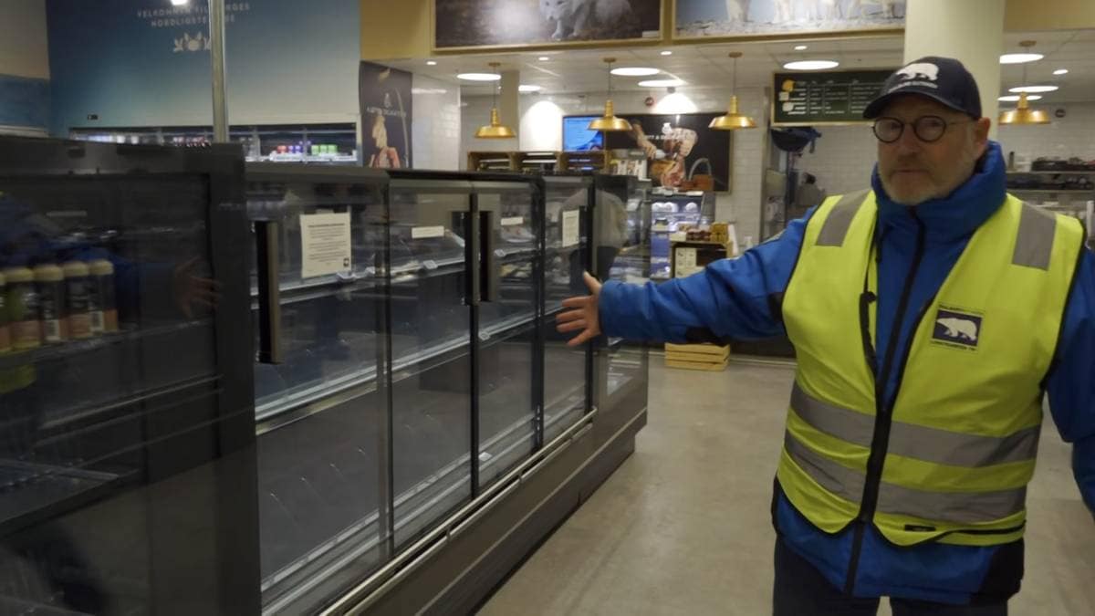 En ansatt i en butikk står foran tomme kjøleskapseksjoner og gestikulerer med hånden. Butikken har en moderne innredning med belysning og hyller i bakgrunnen. Det er flere produkter synlige i de andre kjøleskapene til venstre. En plakat på veggen med bilder av dyr kan sees i bakgrunnen.