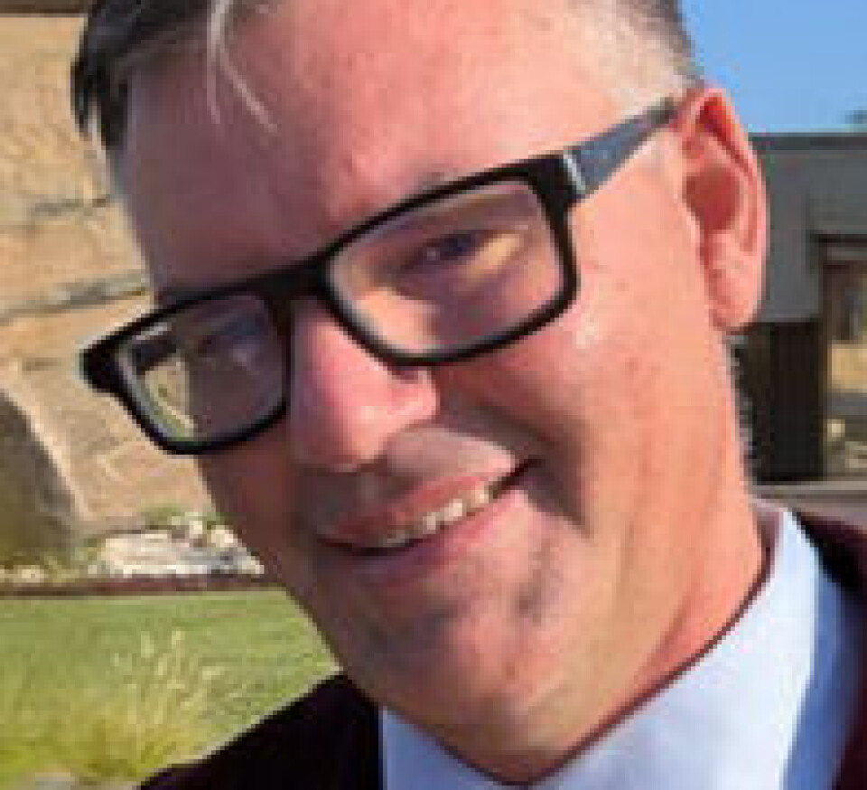 Mann med sorte briller og dressjakke står ute foran en bygning i solskinn.