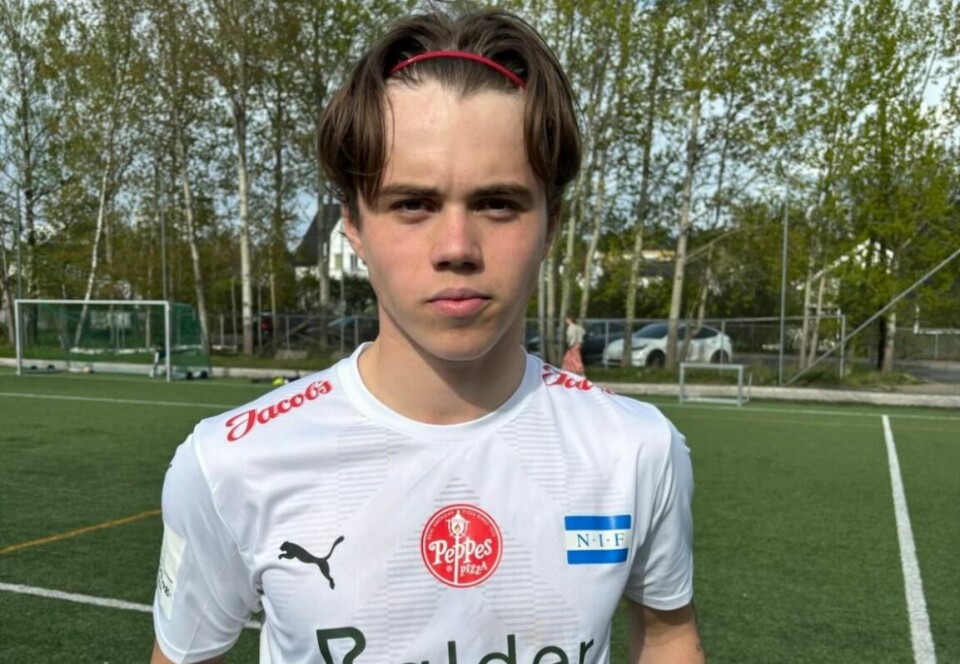 Fotballspiller i hvit drakt står på kunstgressbane med trær i bakgrunnen