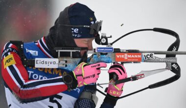 En skiskytter i teknisk utstyr sikter med riflen sin, klart fokusert på målet. Utøveren har på seg en jakke med nasjonsfarger og hansker i sterke farger. Snø faller lett i bakgrunnen, som gir kontekst til utendørsarrangementet. Nummeret "5" er synlig på utøverens rygg. (Bildebeskrivelsen er laget av en KI-tjeneste)