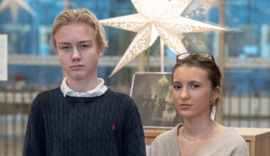 To personer står foran en utstilling i et moderniserte rom. Den første personen har på seg en mørk genser og jeans, mens den andre har en lys cardigan og mørke bukser. Bak dem ses en dekorativ stjerne i taket og informasjonsplakater på veggen. Rommet har store vinduer og et minimalistisk design. (Bildebeskrivelsen er laget av en KI-tjeneste)