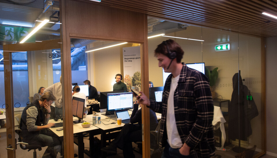 Åpent kontor med unge ansatte foran skjermer og en person i telefon i glasskorridor.